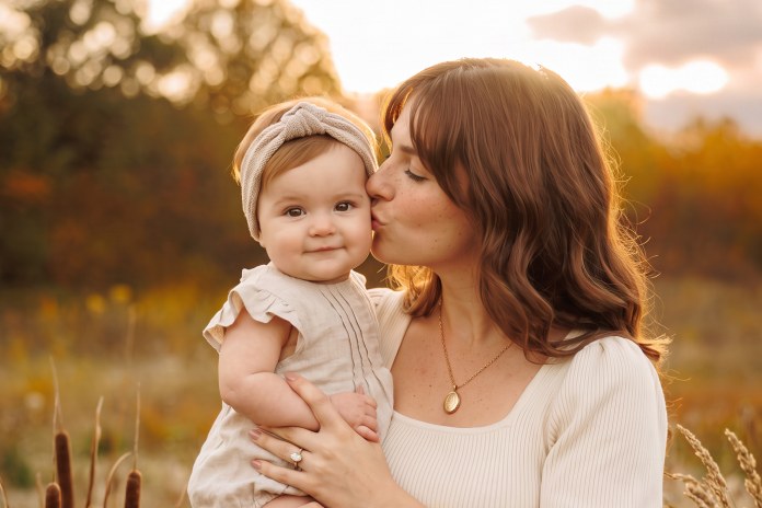 Parent kissing their baby on the cheek in a field at sunset