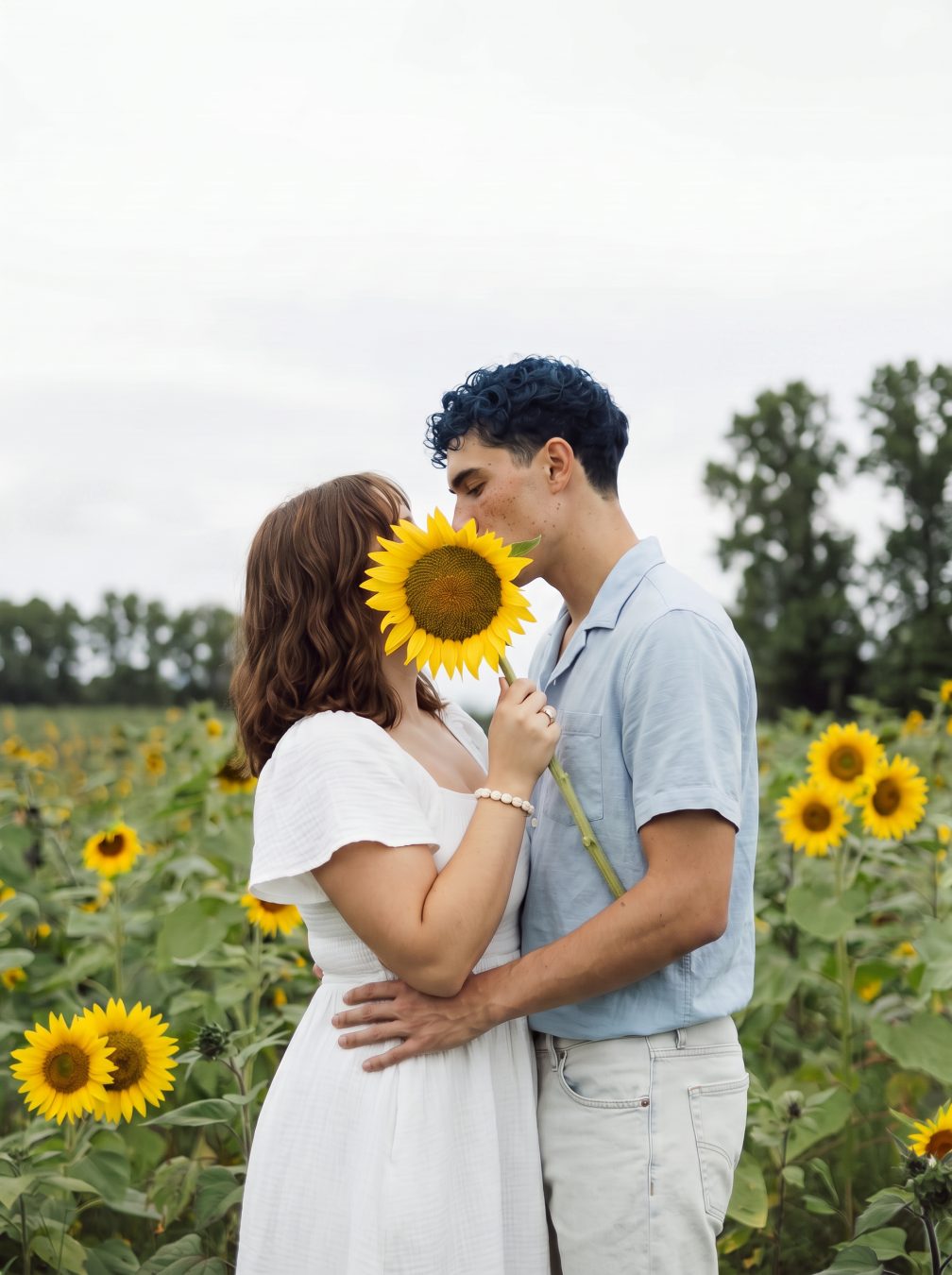 Couple in a sunflower field, one holding a sunflower in front of their face
